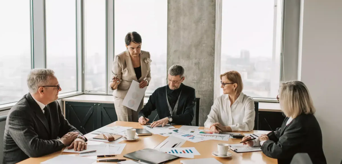 A group of business professionals engaged in a discussion around a conference table in a modern meeting room.